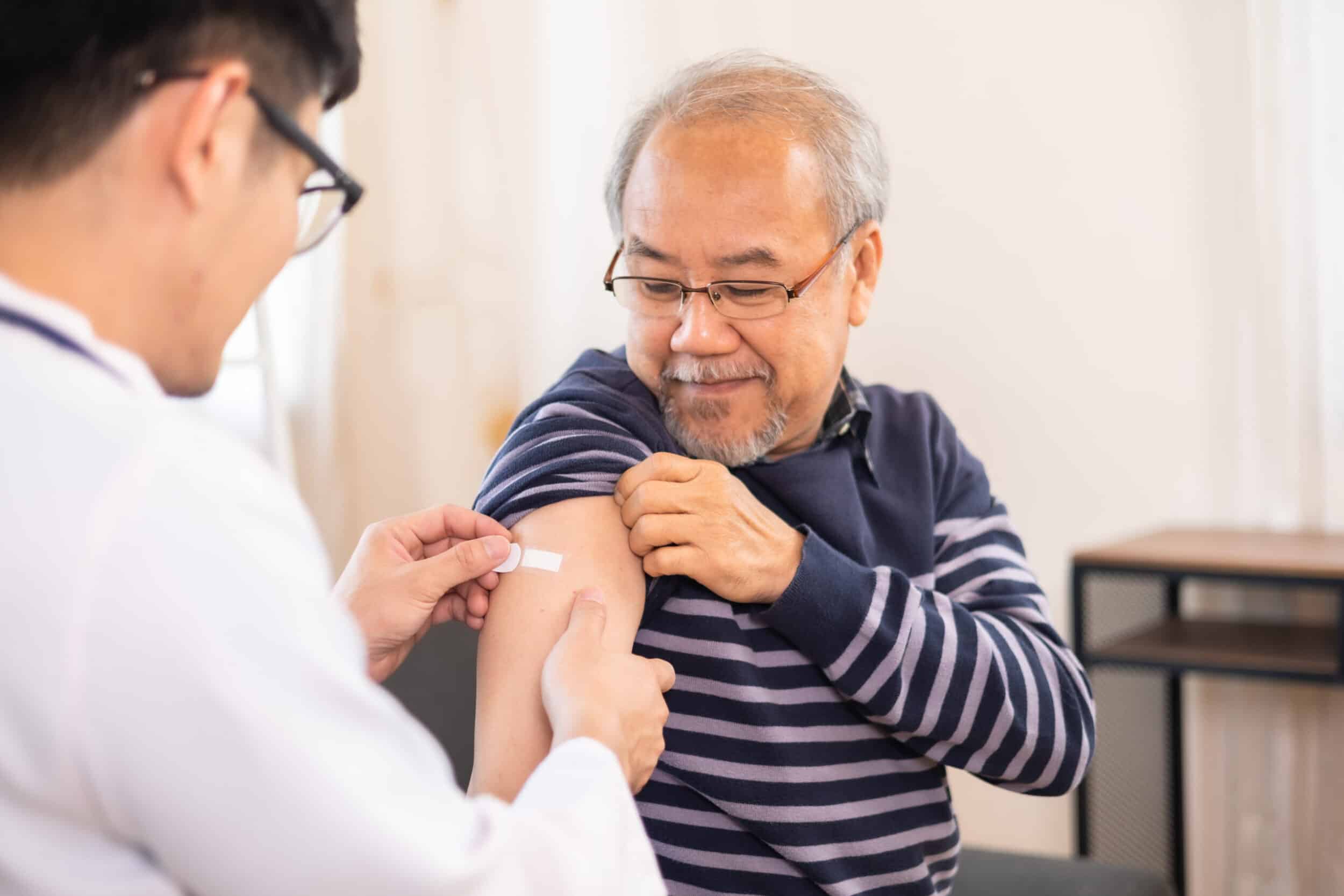 Safe vaccination for old people. Elder man in medical getting flu or Covid 19 vaccine sitting on sofa at home. Asian Doctor or nurse giving flu or Covid 19 shot to senior.